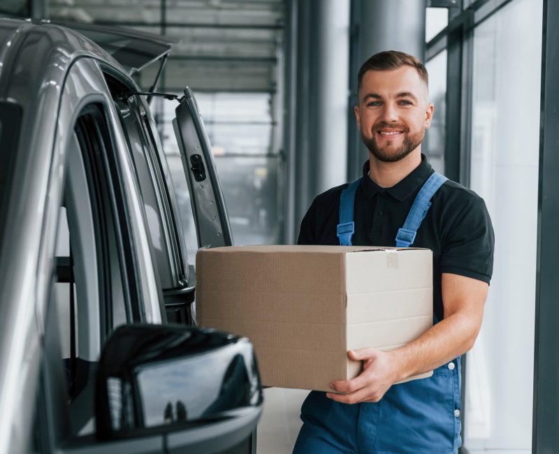 With box in hands. Delivery man in uniform is indoors with car and with order